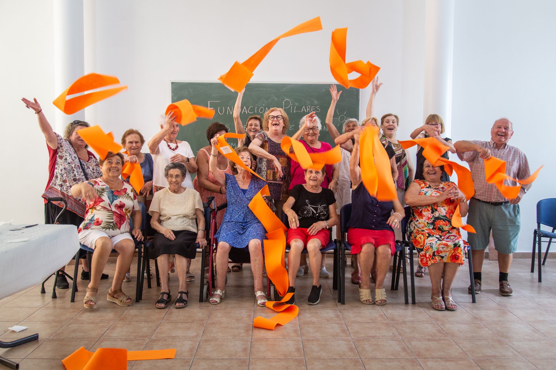 Grupo de personas participando en una actividad grupal en un aula, lanzando cintas naranjas al aire como parte de un taller organizado por Fundación Pilares